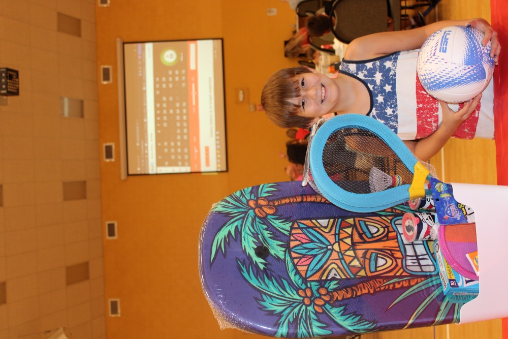 Smiling boy in a red, white, and blue tank top stands by a prize basket filled with beach toys and holds a white ESPN ball at the South Cape Community Center family bingo night.
